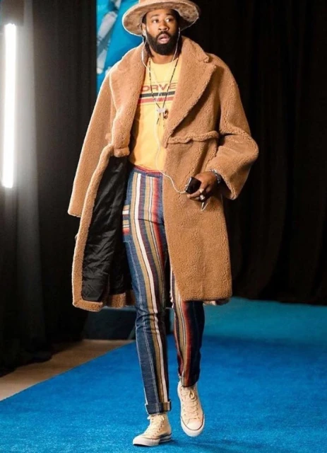 A bearded man wearing a hat and a tan sherpa coat walks on a blue carpet. He is wearing yellow shirt, striped pants, and cream-colored sneakers.
