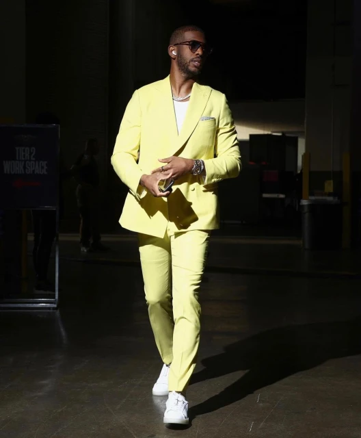 Adult male walks through a dark corridor wearing a yellow suit, white shirt, and white sneakers. He also wears sunglasses and jewelry.