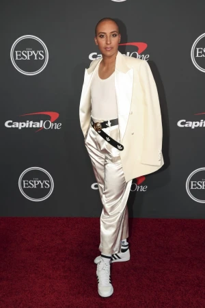 A person with a shaved head poses on a red carpet in a white suit and matching shoes at the ESPYS award show, smiling at the camera.