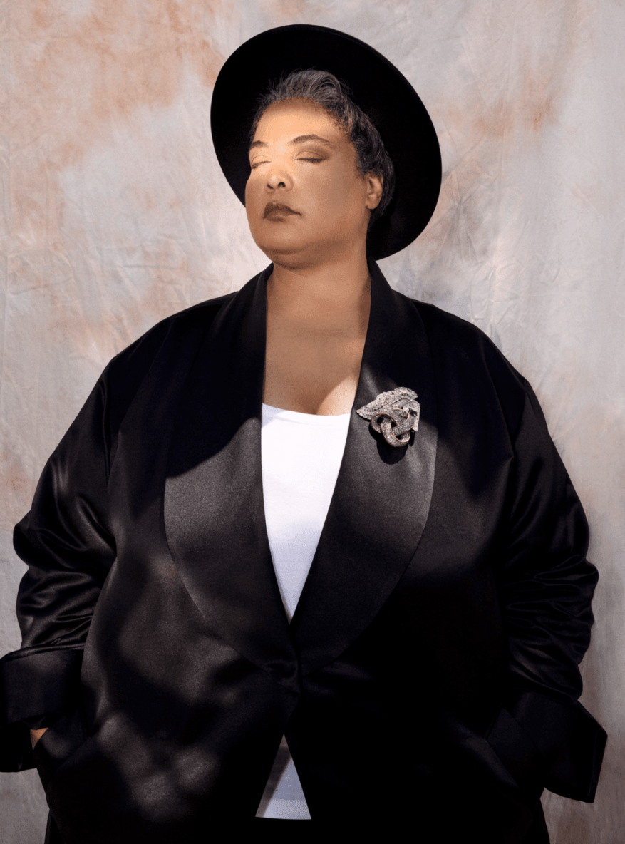 Portrait of an African-American woman wearing a black hat and jacket, adorned with a jeweled panther brooch, eyes closed in warm lighting.