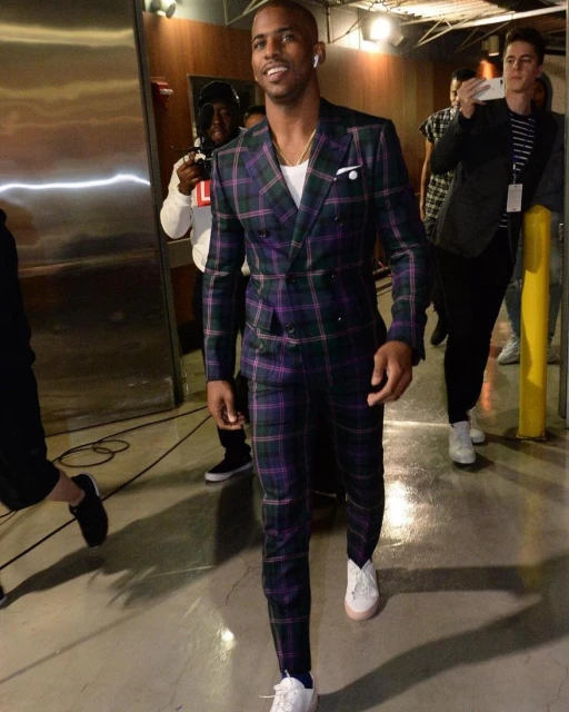 Chris Paul walks in a green and purple plaid suit, wearing white sneakers, smiling slightly with a few other people in the background.