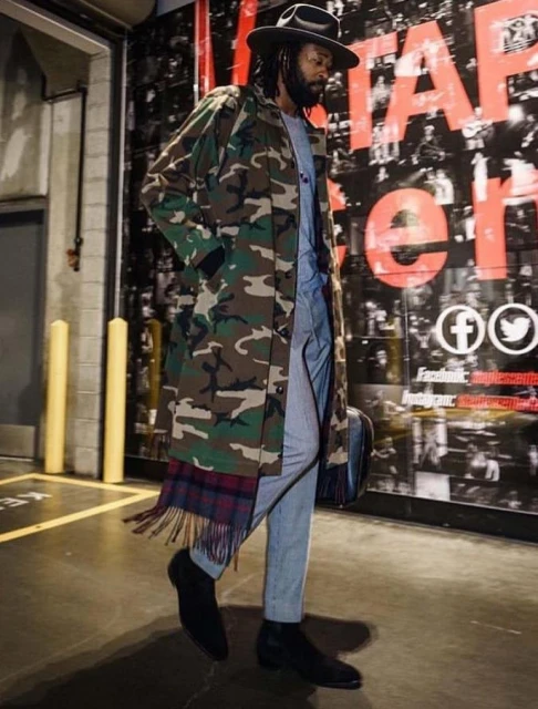 Man in camo trench coat, fedora, and grey suit walks indoors with patterned scarf; social media icons on background wall.