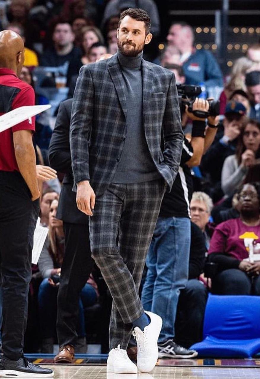 Well-dressed man in a grey plaid suit on a basketball court, standing with legs crossed, with blurred spectators and cameramen in background.
