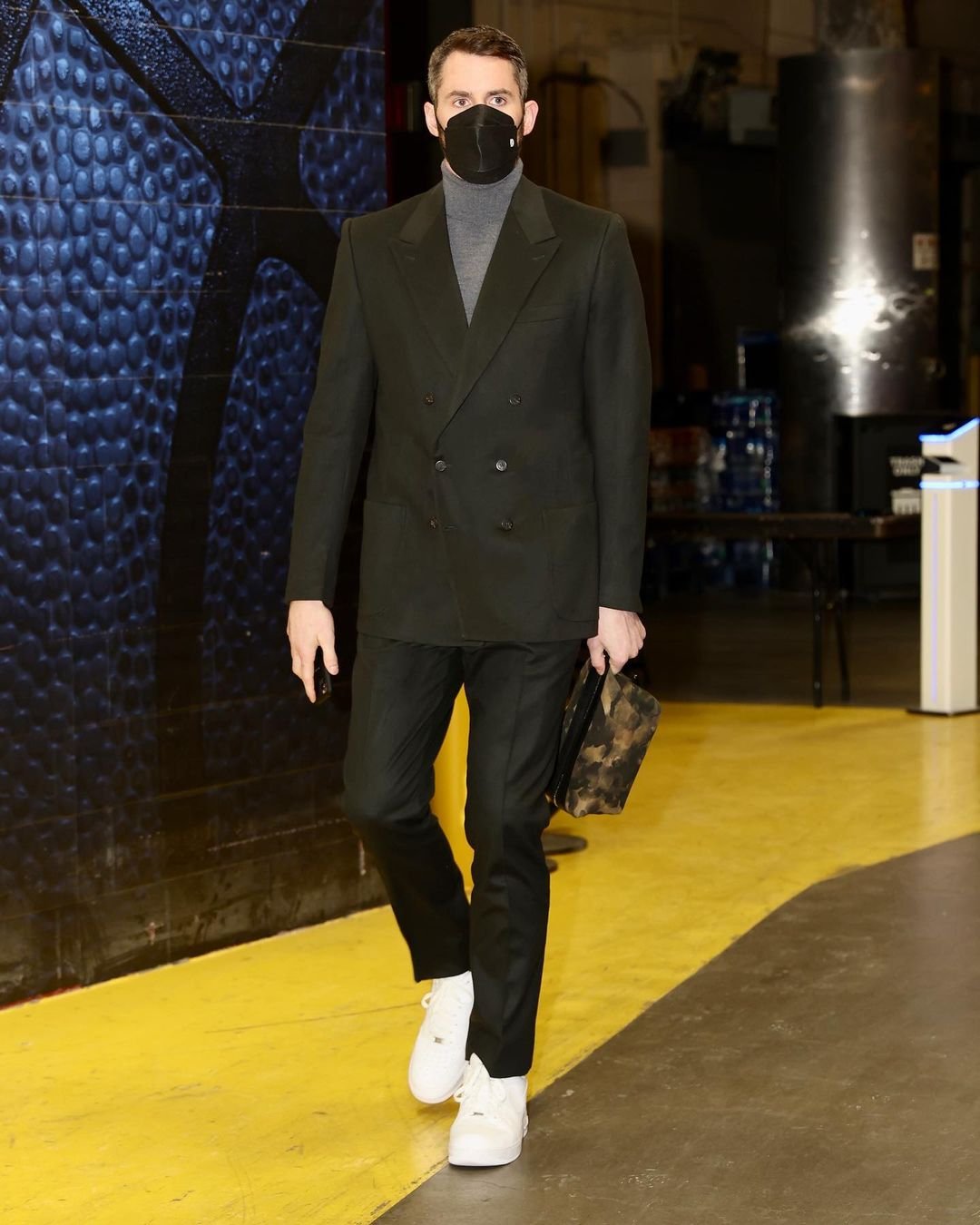 Man in black coat and grey turtleneck, wearing a face mask, carrying a briefcase, walking on yellow marked floor.