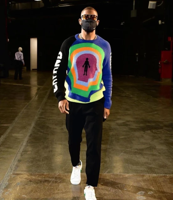 An adult male in sunglasses and a face mask walks indoors wearing a colorful graphic sweater, dark pants, and white sneakers.
