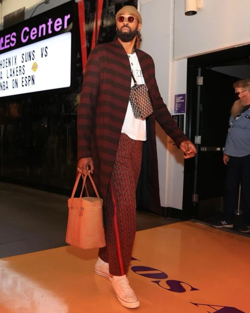 Stylish man in sunglasses walks on a basketball court wearing a striped coat, patterned pants, carrying a handbag, and accessorized.