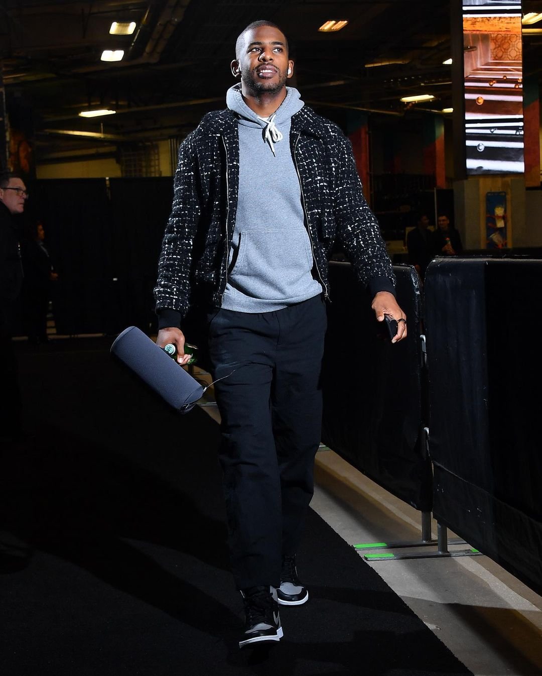 Adult male in a patterned black jacket and grey hoodie walks indoors holding a rolled object, showcasing a casual yet stylish look.