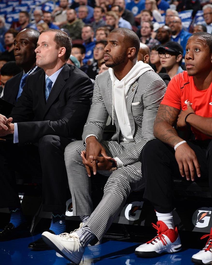 Three adult males sit on a bench in a stadium, with one wearing a houndstooth suit and white hoodie, and a crowd in the background.
