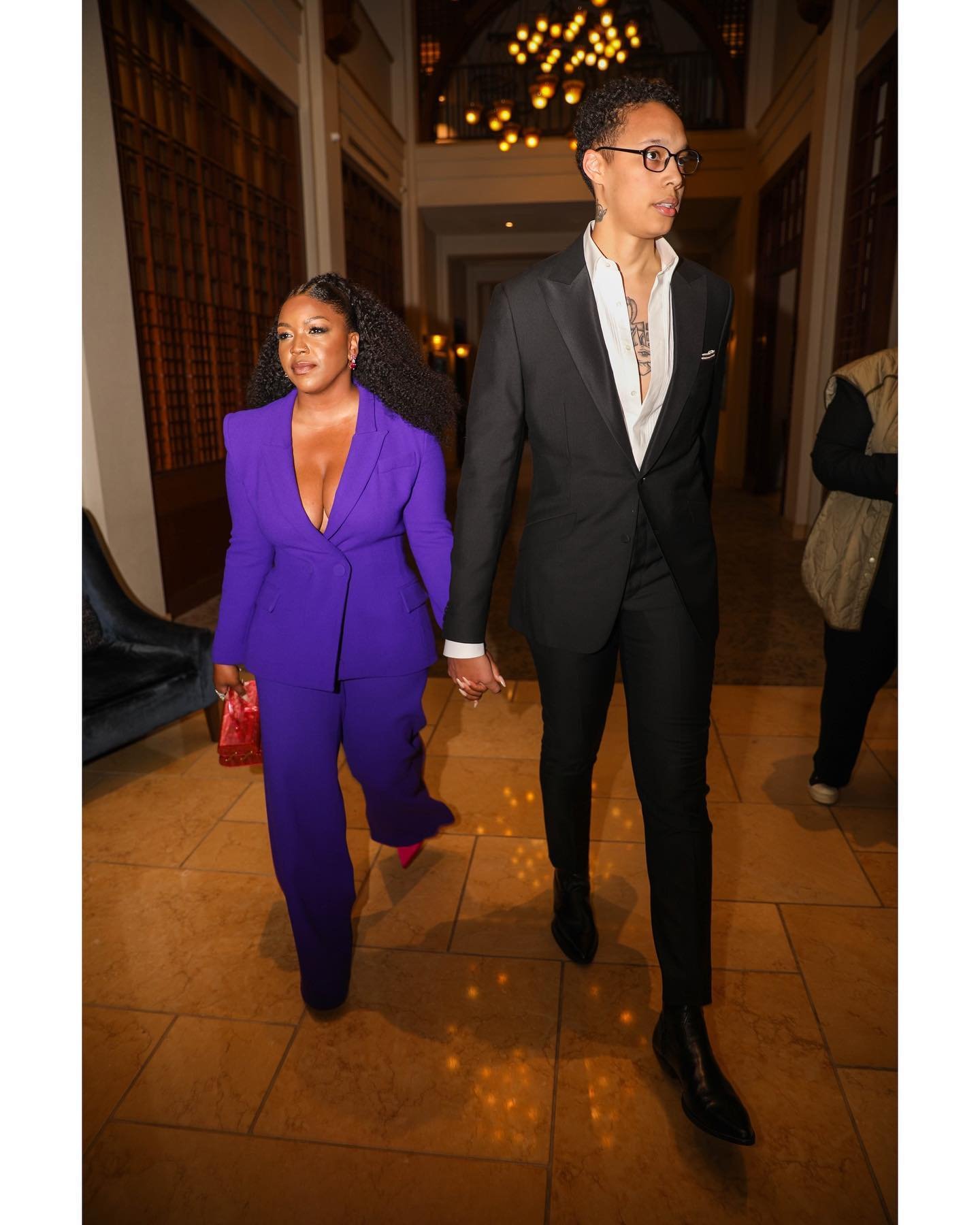 Two women in a corridor, one wearing a purple suit and the other in a black suit, holding hands. Interior with neutral tones.