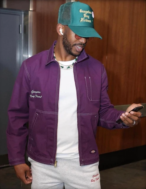 Man in purple jacket, white tee, and grey sweatpants, wearing a green trucker hat and sunglasses, holding a smartphone indoors.