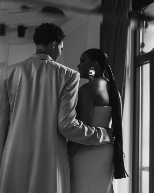 Black and white shot of a mixed-race couple indoors: a man in a jacket and a woman in a dress embracing by the window.