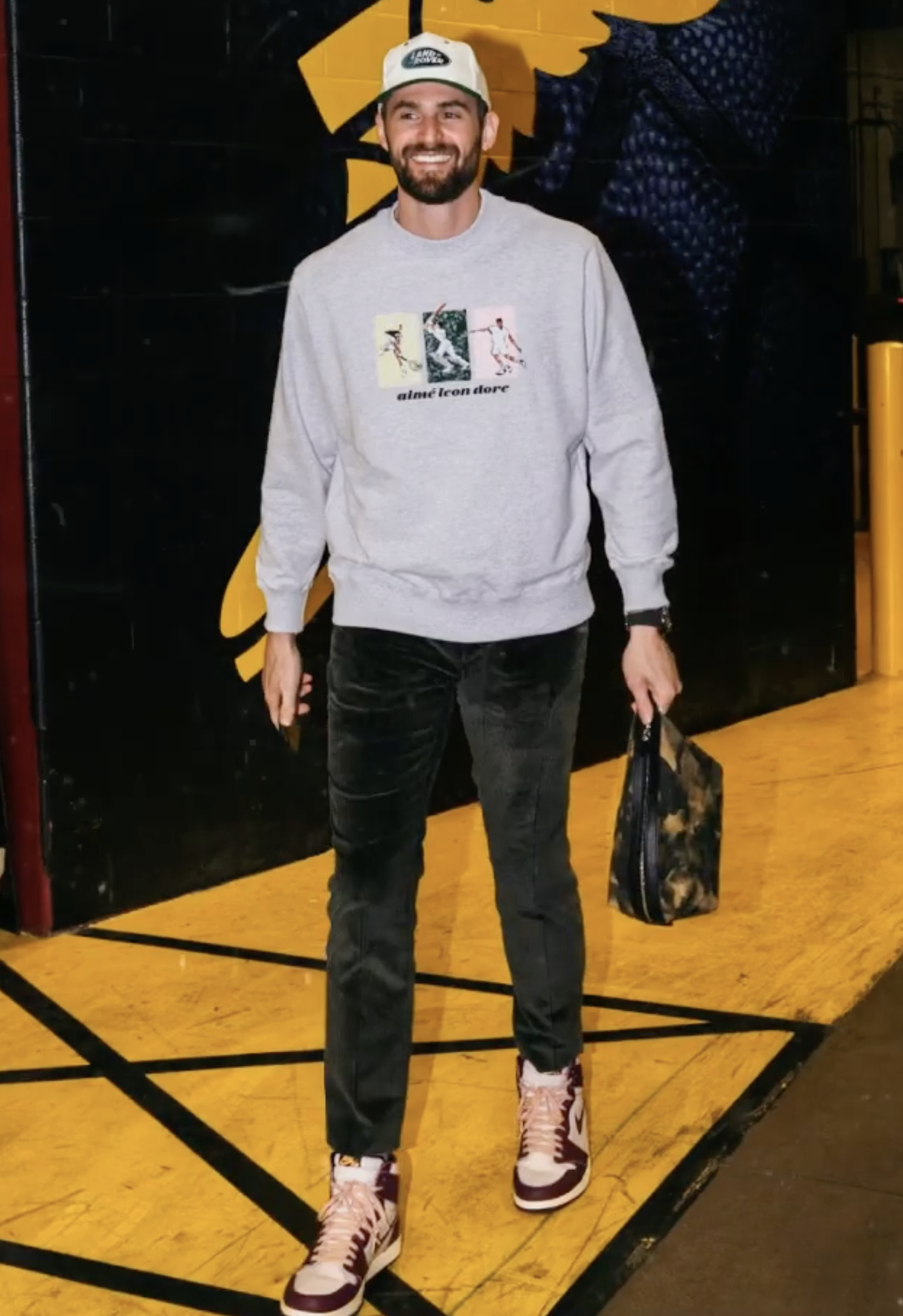 Adult male in a light gray sweatshirt with colorful graphics paired with dark green pants, baseball cap, and sneakers. 