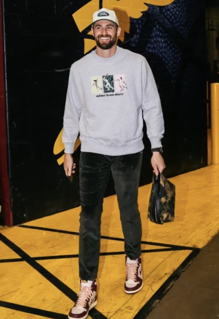 Adult male in a light gray sweatshirt with colorful graphics paired with dark green pants, baseball cap, and sneakers. 