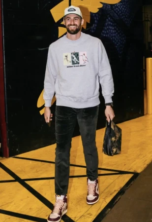 Adult male in a light gray sweatshirt with colorful graphics paired with dark green pants, baseball cap, and sneakers. 