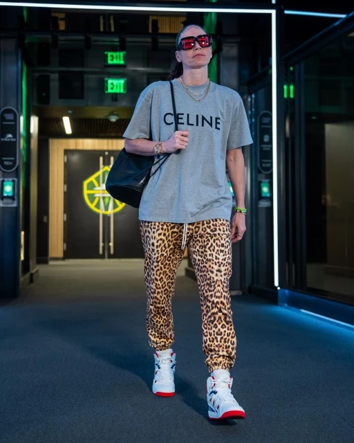 Adult female poses in leopard print pants and a grey tee in an indoor hallway with directional lighting and white high-top sneakers.