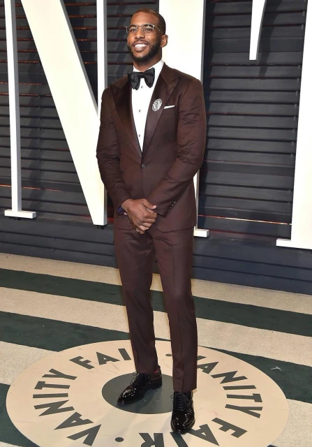 Full-length shot of Chris Paul in a brown suit with a bow tie, standing indoors on a Vanity Fair-branded surface.