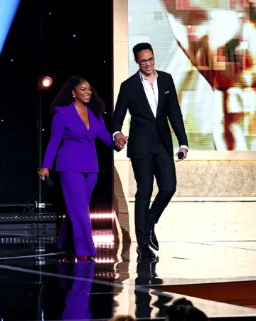 Two presenters, an adult woman in a purple suit and an adult man in a black suit, walk on stage during an awards show event.