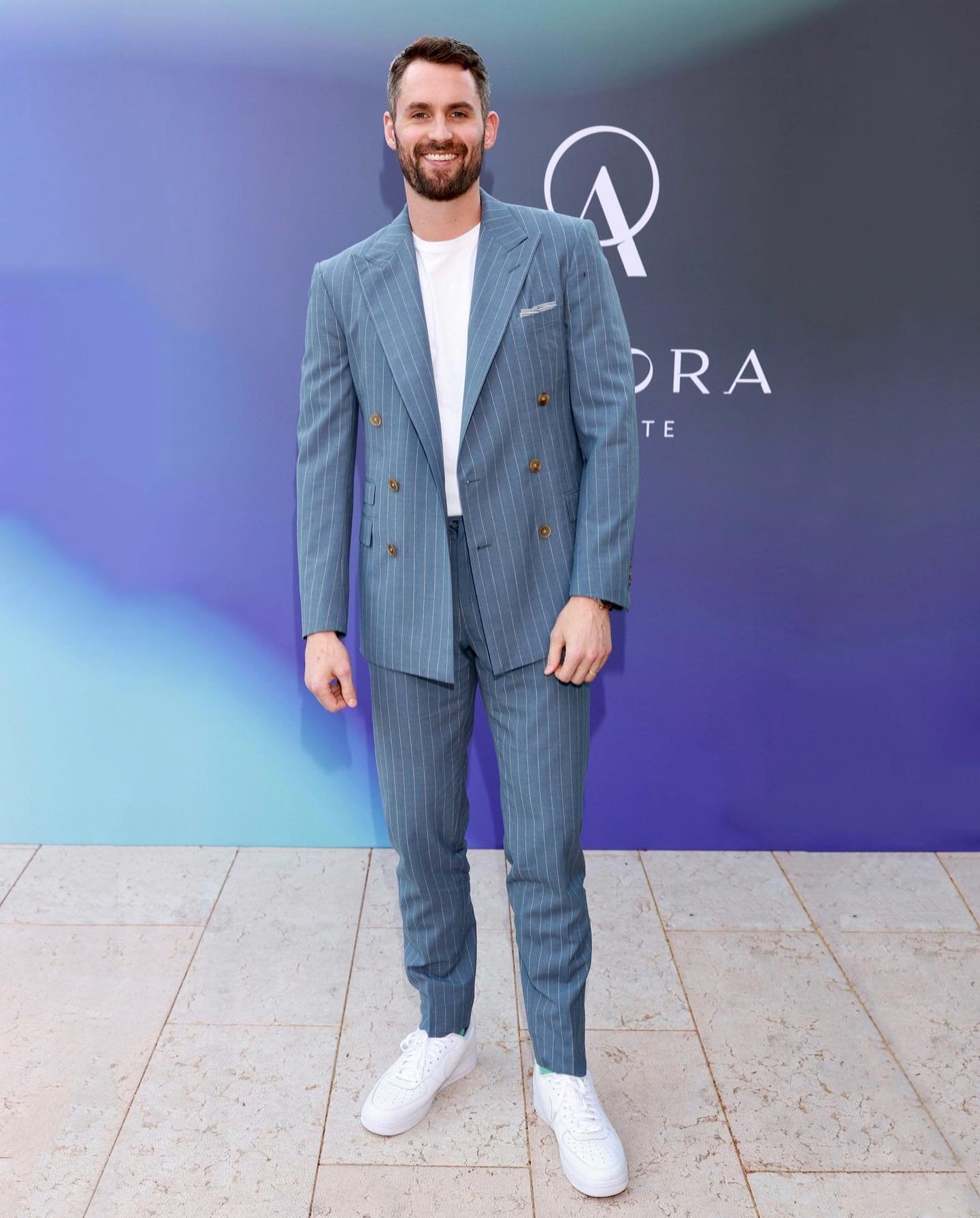 Adult man in a pinstripe suit with white sneakers posing outdoors against a blue and purple backdrop with architectural details.
