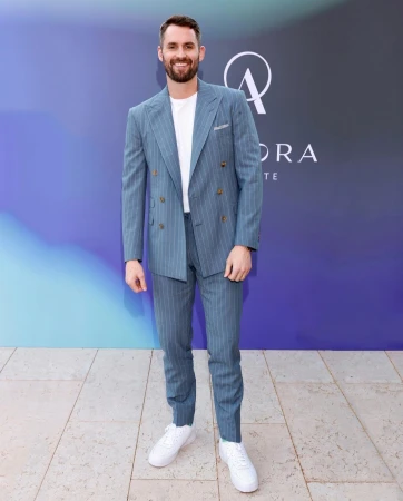 Adult man in a pinstripe suit with white sneakers posing outdoors against a blue and purple backdrop with architectural details.