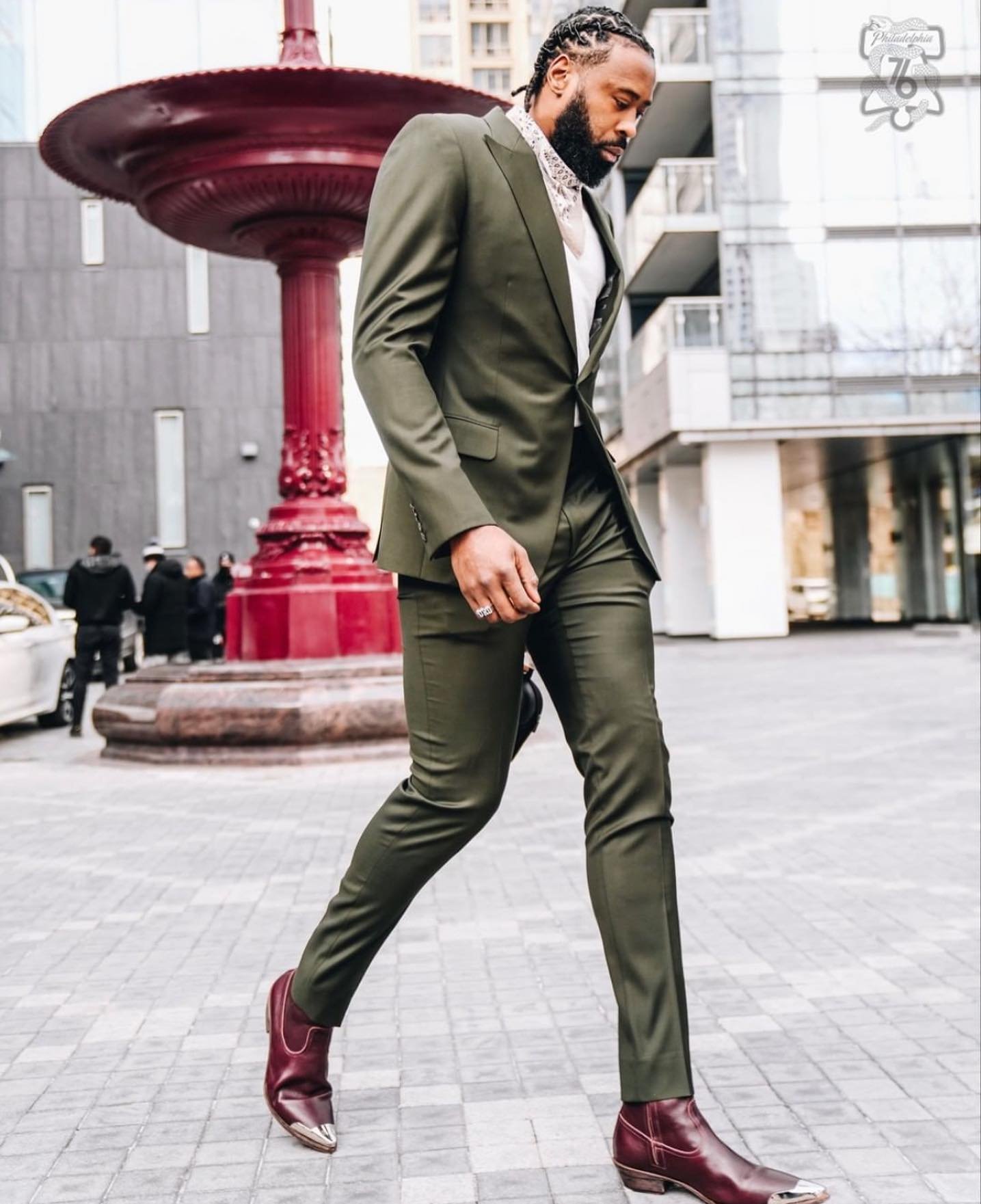 A man in a green suit walks on a paved street in front of a decorative fountain, showcasing a sharp, stylish, and urban look.