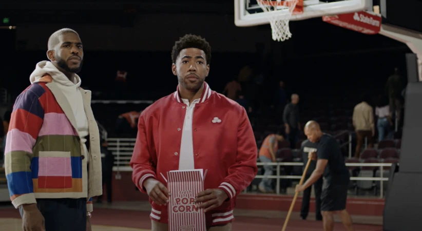 Two adult males stand on a basketball court; one in a red jacket holds a popcorn bag while the other wears a colorful jacket.