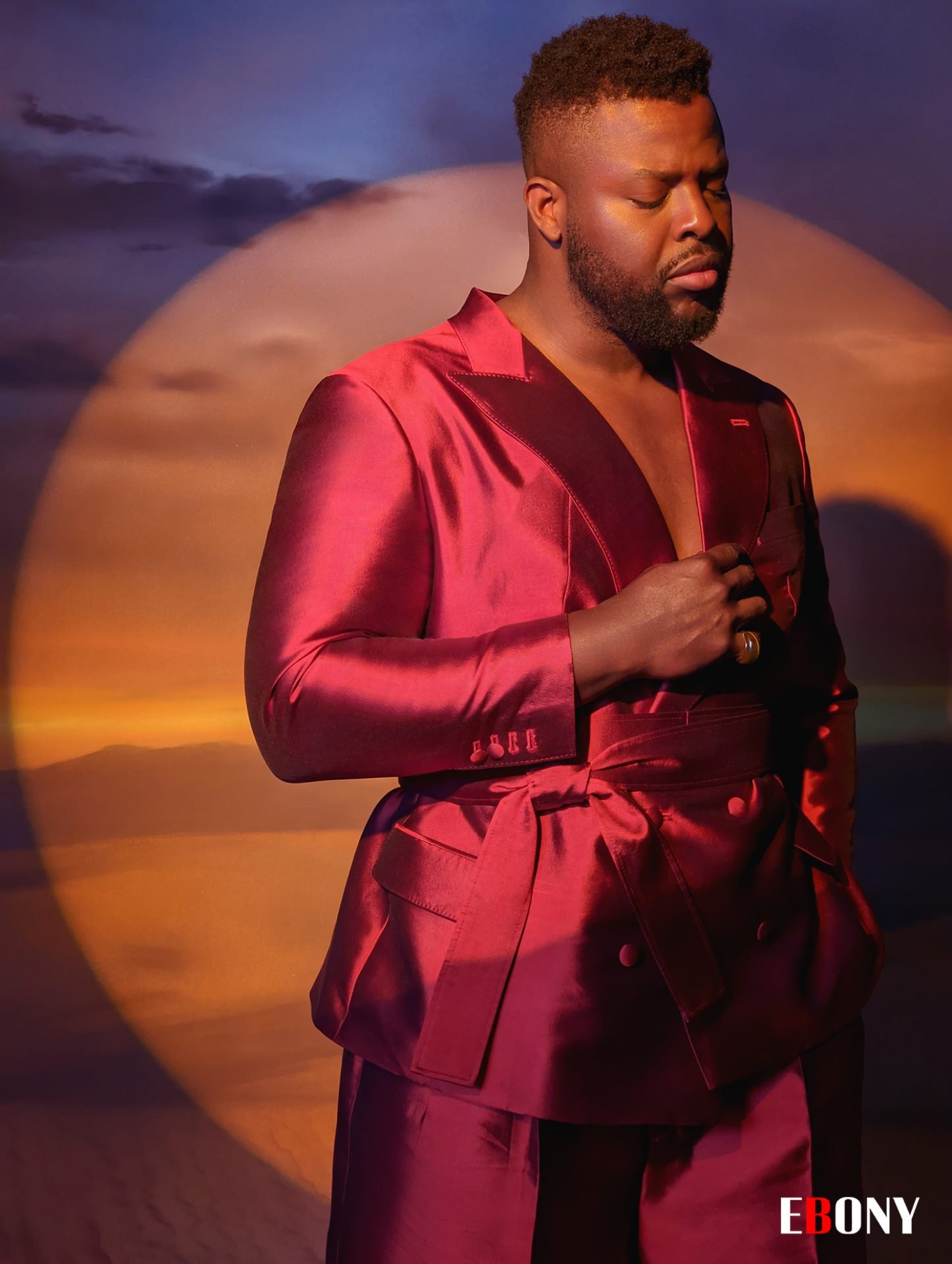 Man in a maroon suit standing indoors with a spotlight effect. Features high-fashion attire, male model, and professional studio lighting.