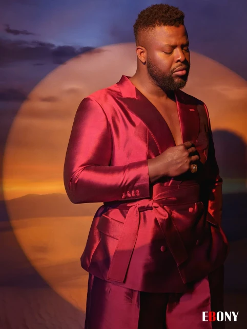 Man in a maroon suit standing indoors with a spotlight effect. Features high-fashion attire, male model, and professional studio lighting.
