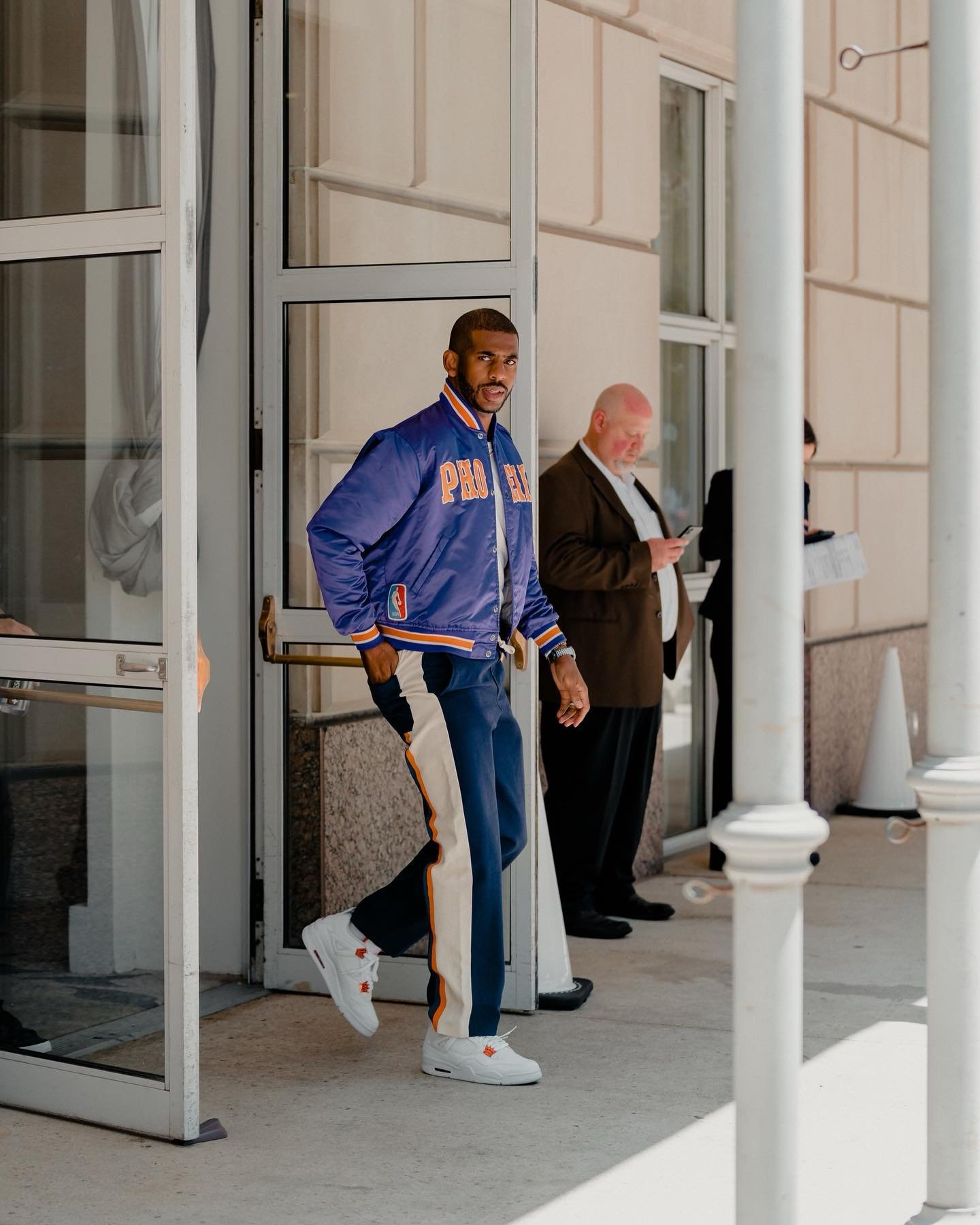 A man wearing a blue jacket and track pants walks out of a building, while a man holding a phone stands in the background.