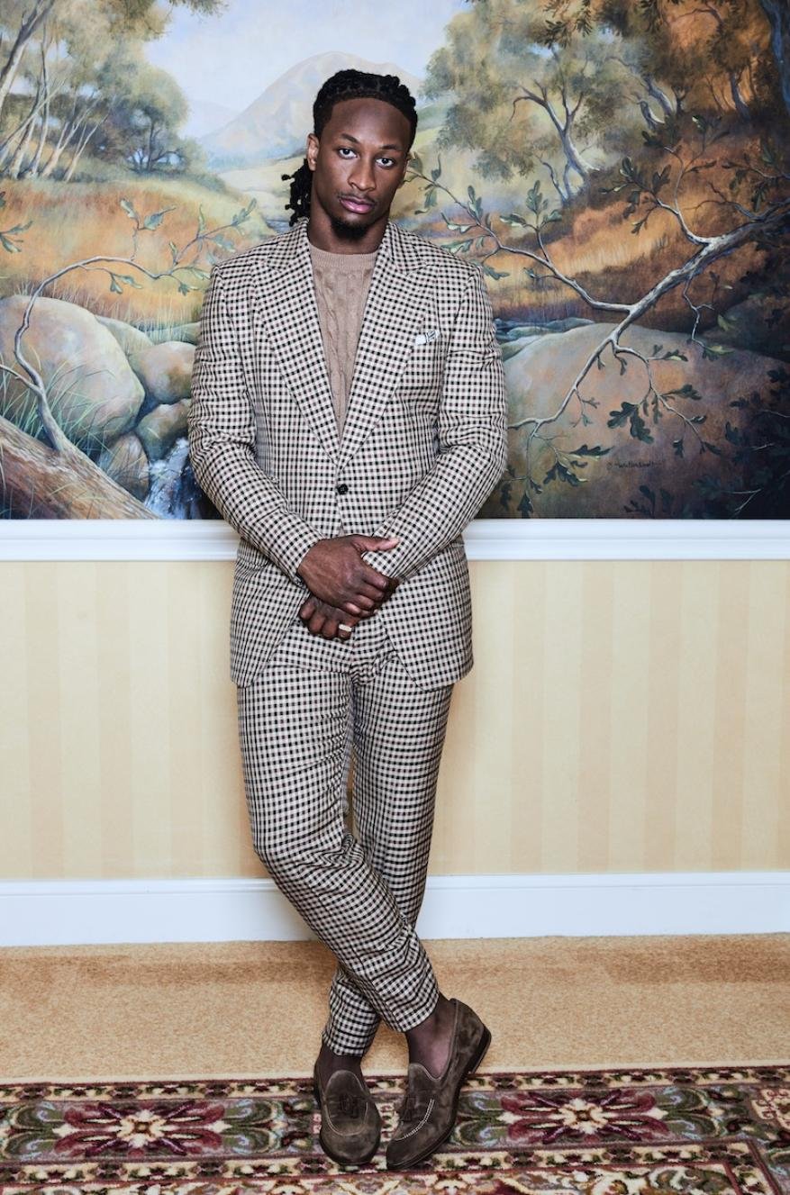 Man in a beige and brown plaid suit standing in front of a landscape painting. He is wearing brown loafers and has his legs crossed.