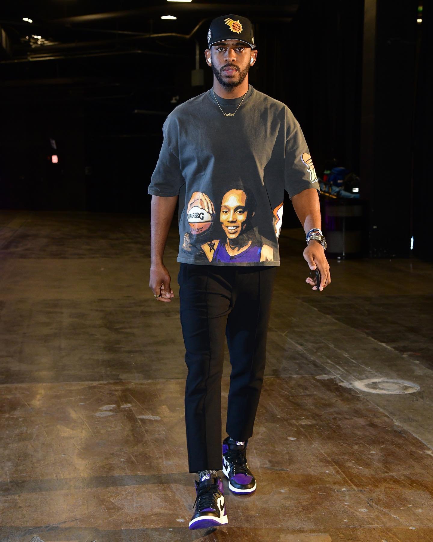 Male model in a grey T-shirt with an image and black pants. High-top purple and black sneakers, and baseball cap in a studio-like setting.