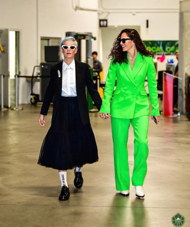 Two women walking hand-in-hand through a corridor, one in a green suit and the other in a black and white ensemble.