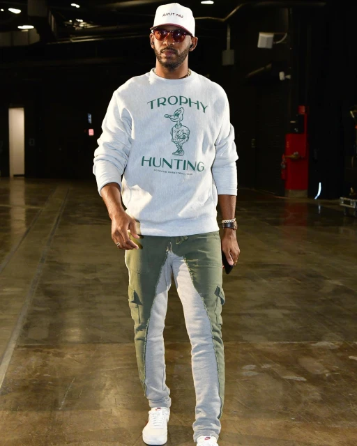 Man in casual fashion stands indoors wearing a sweatshirt, two-tone jeans, sneakers, and a baseball cap, with a dark background.