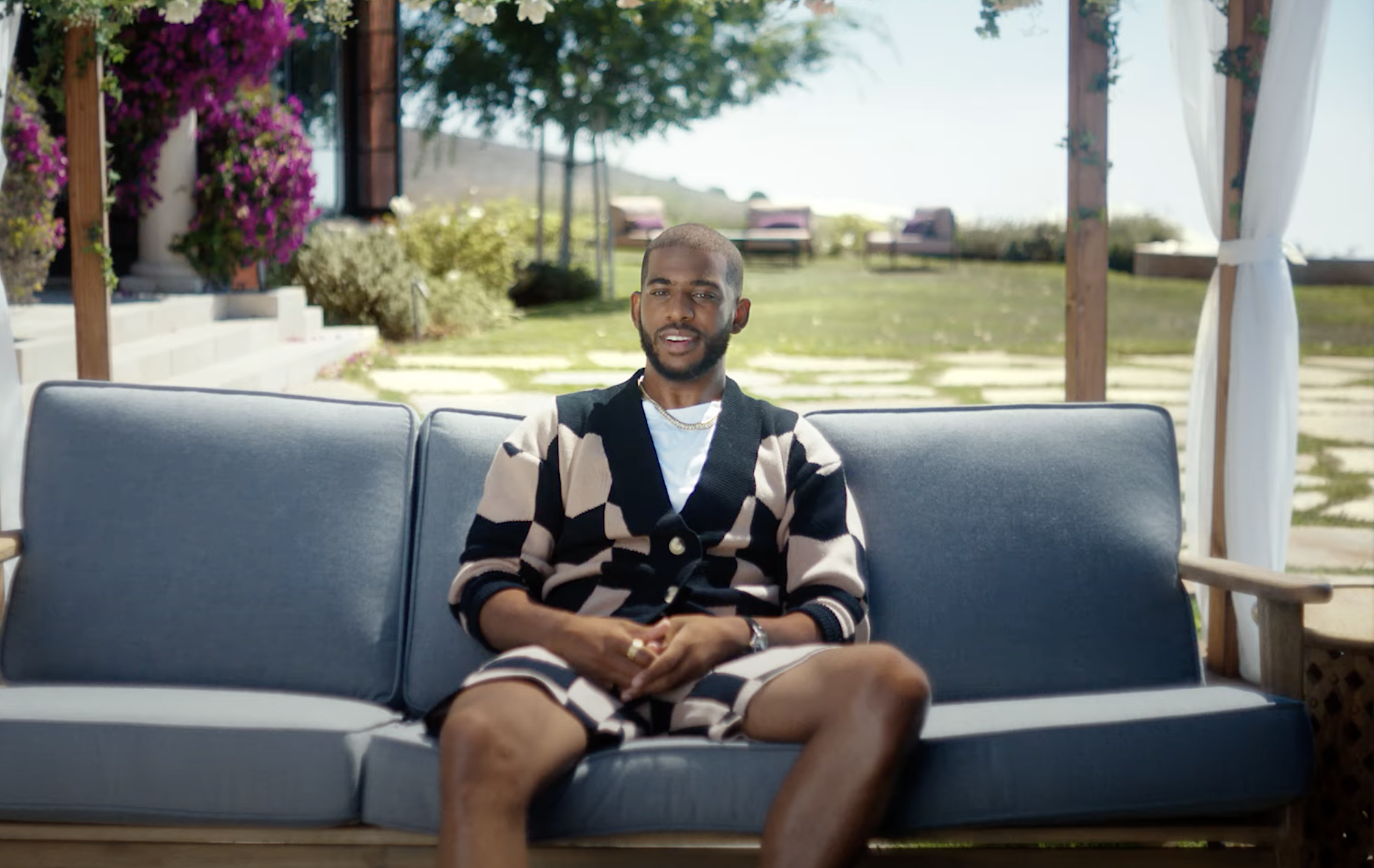 An adult man sits on an outdoor couch in a garden setting. The man wears a patterned cardigan and shorts, and smiles towards the camera.