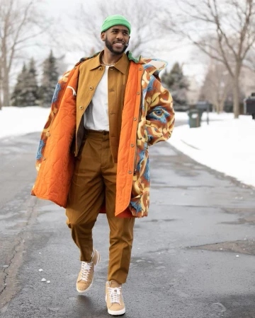 Man in brown suit and patterned coat walking on road in snowy outdoor setting, wearing green beanie, with natural light.