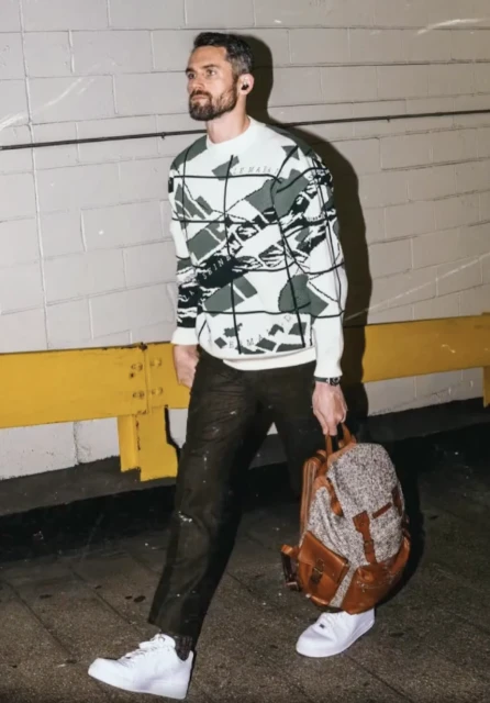 An adult male is walking in an urban setting, carrying a backpack and wearing a patterned sweater, dark pants, and white sneakers.