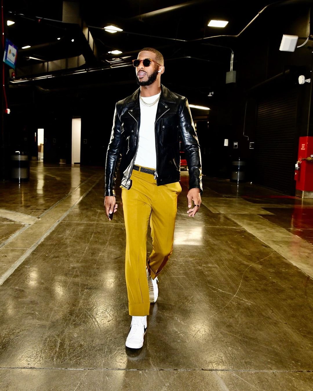 Stylish adult man walking in a tunnel, wearing a black leather jacket, mustard-colored pants, sunglasses, and white sneakers in indoor setting.