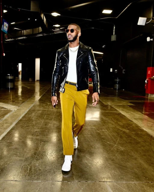 Stylish adult man walking in a tunnel, wearing a black leather jacket, mustard-colored pants, sunglasses, and white sneakers in indoor setting.