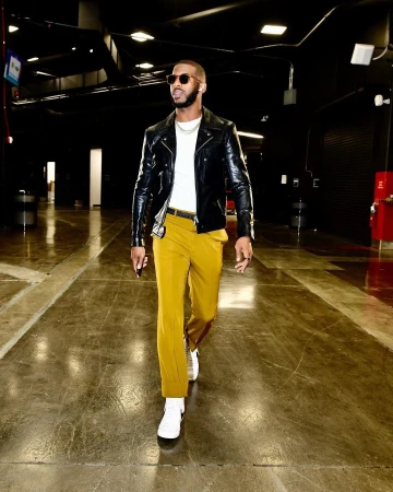 Stylish adult man walking in a tunnel, wearing a black leather jacket, mustard-colored pants, sunglasses, and white sneakers in indoor setting.