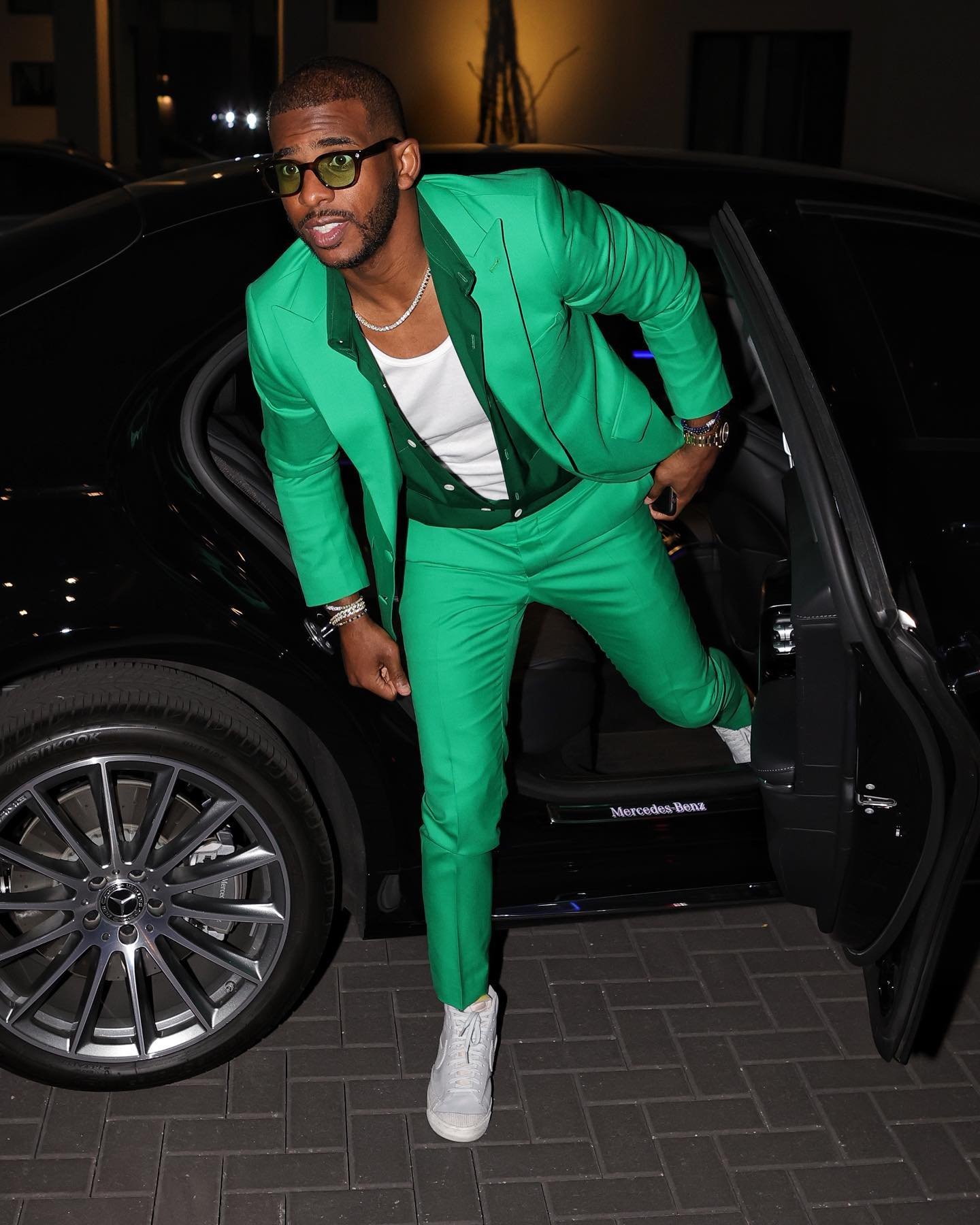 Man in a green suit stepping out of a black Mercedes-Benz, wearing glasses and white sneakers, posing for a photograph at night.