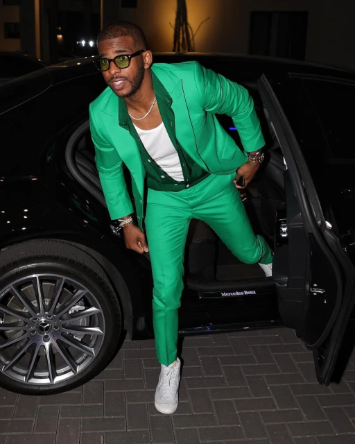 Man in a green suit stepping out of a black Mercedes-Benz, wearing glasses and white sneakers, posing for a photograph at night.