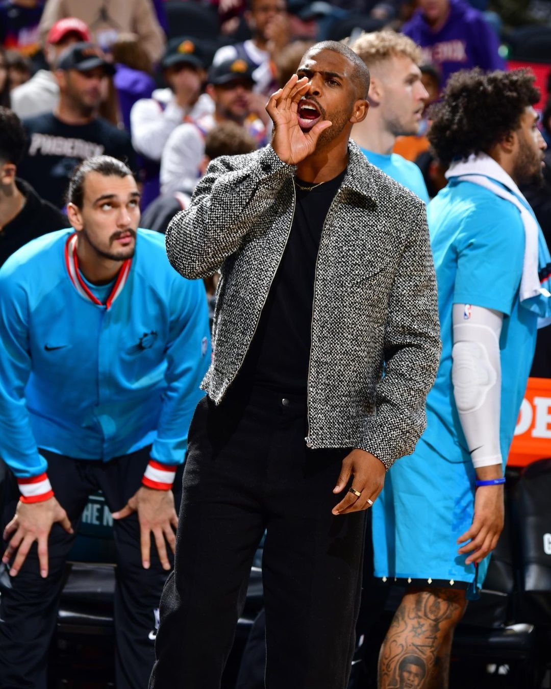 Chris Paul courtside at a basketball game, dressed in a tweed jacket, black pants, and black shirt, calling out to the players. 