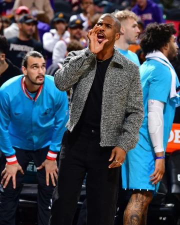 Chris Paul courtside at a basketball game, dressed in a tweed jacket, black pants, and black shirt, calling out to the players. 