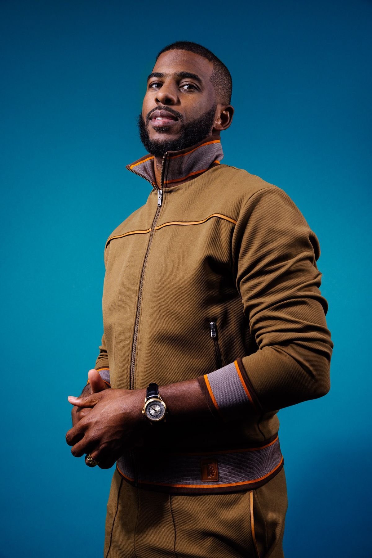 Portrait of an adult male wearing a brown tracksuit with a turquoise background. He is looking at the camera with a neutral facial expression.