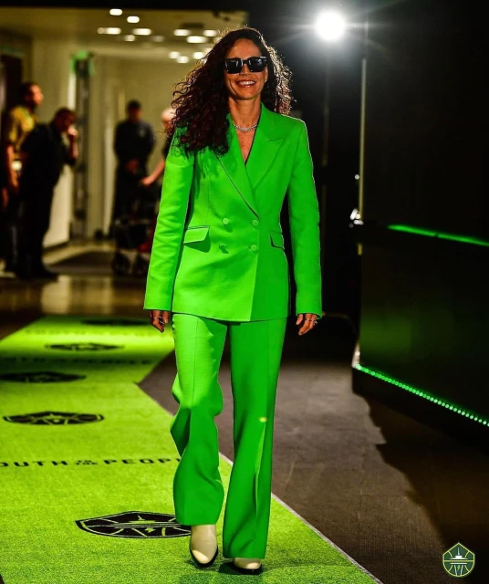 A woman in a bright green suit walks down a green carpeted walkway, wearing sunglasses, with blurred figures in the background.