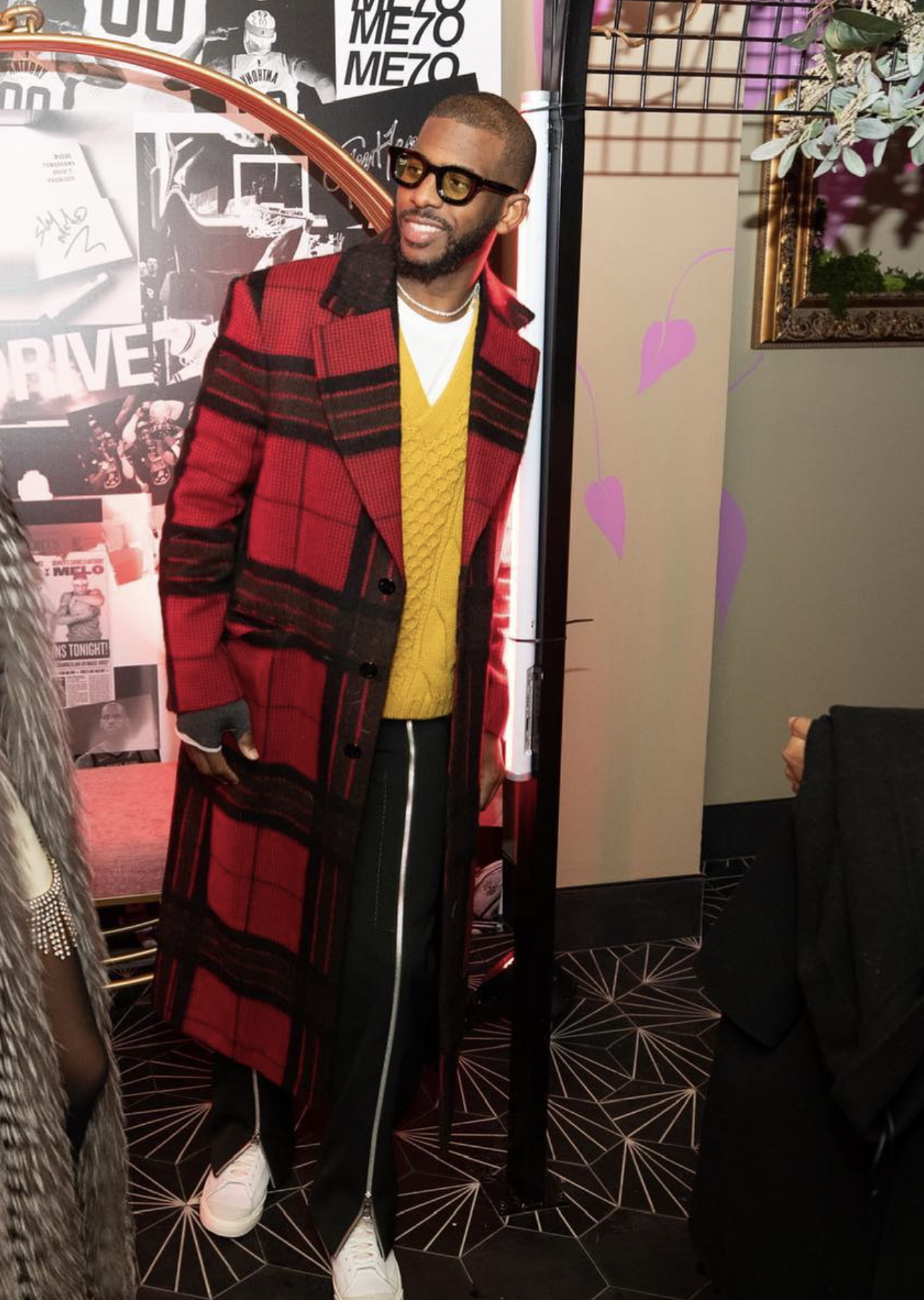 A man in a red plaid coat, yellow sweater, and white sneakers poses in an indoor setting with modern geometric patterned floors. 