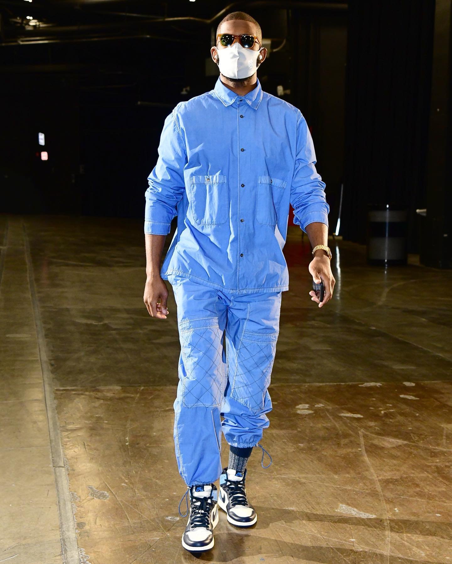 Stylish man in blue outfit and sneakers walking indoors with a white face mask, wrist watch, and smartphone against a dark background.