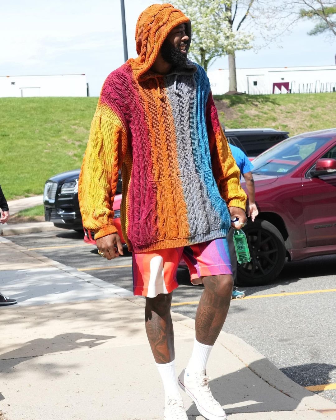 Adult male wearing colorful hooded sweater and shorts stands on a city street on a sunny day, with cars and buildings in the background.