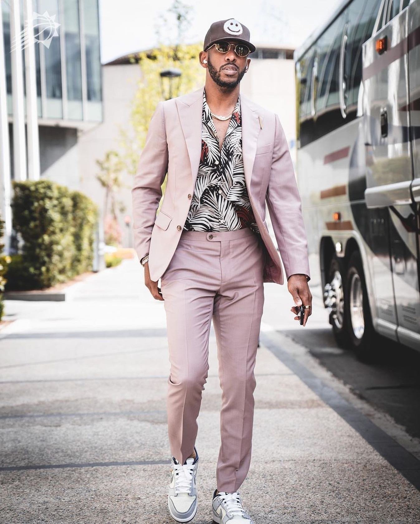Man in a rose-colored suit walks past a bus with a brown smiley face hat and floral shirt, accessorized with a chain necklace.