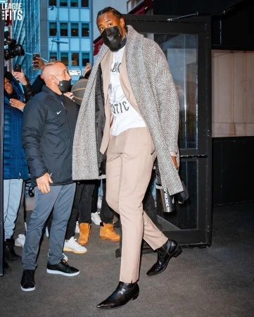 A tall adult male strides out of a building, wearing a long herringbone coat over a tan suit, while people photograph him.