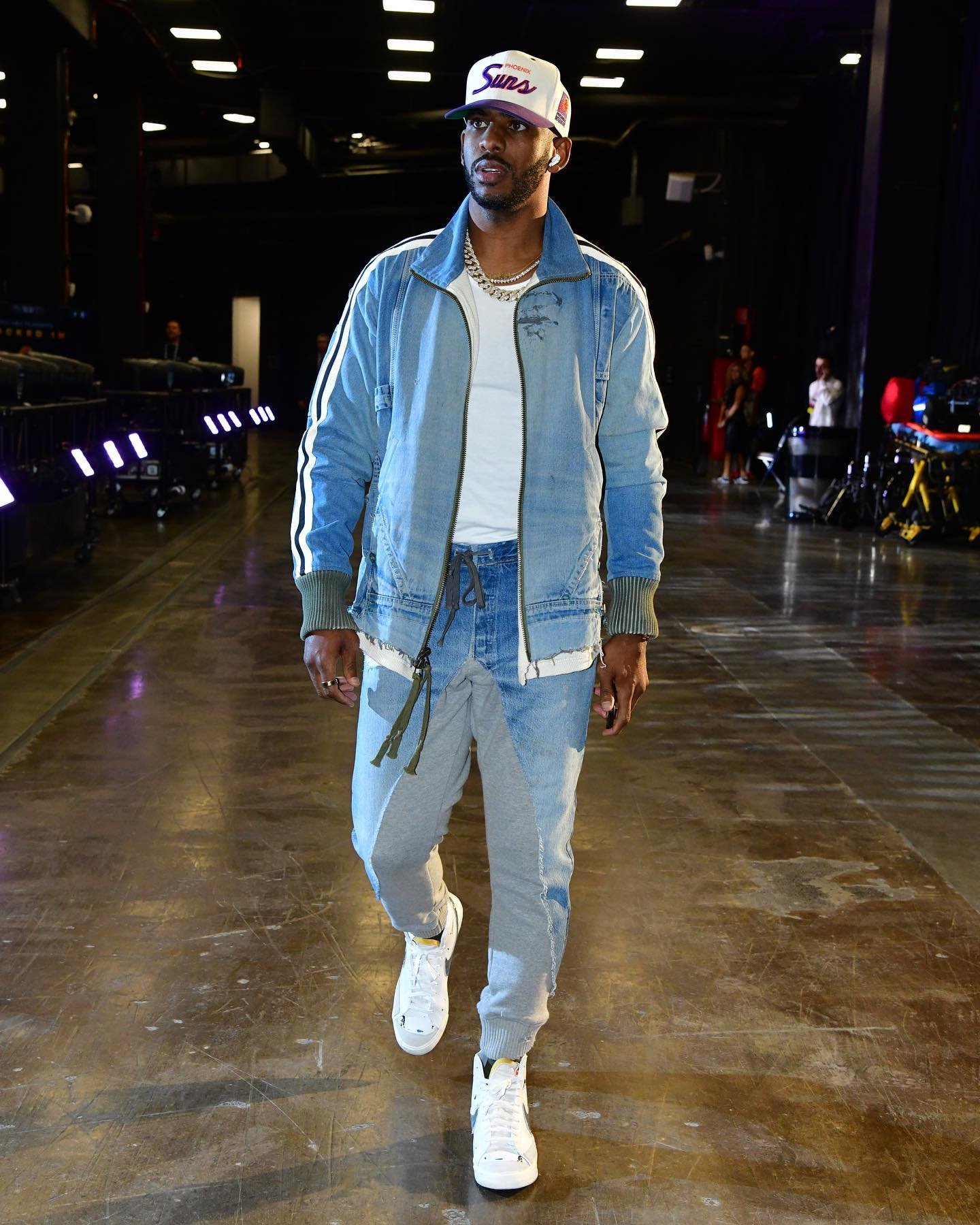 An adult male fashion model wearing a hat, blue jean jacket, t-shirt, and pants, walking on a glossy surface indoors.